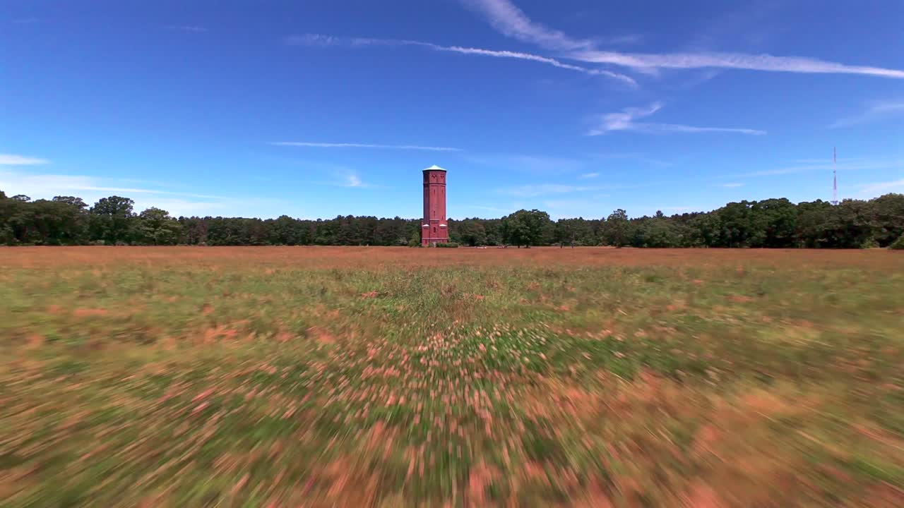 Aerial FPV flight towards a brick water tower on a sunny day in a large grass field on the Pilgrim Psychiatric Center property. The camera dolly in to the structure at a low altitude just above grass