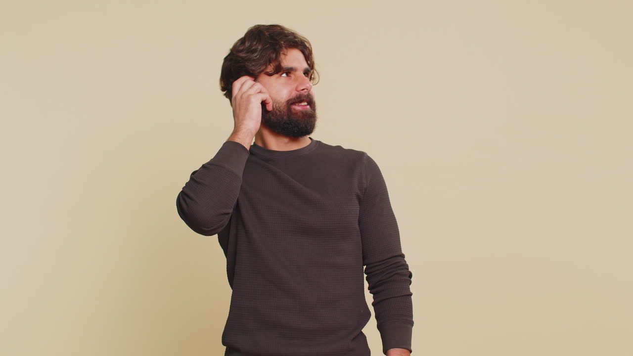 Giovane uomo ispanico pensieroso che guarda di lato con espressione positiva su sfondo da studio beige