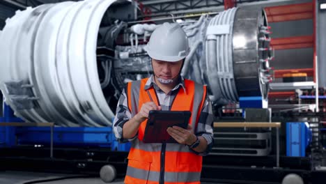 asian male engineer with safety helmet taking note on the tablet and looking around while standing with airplane engine maintenance conducted, analytics checking the turbine