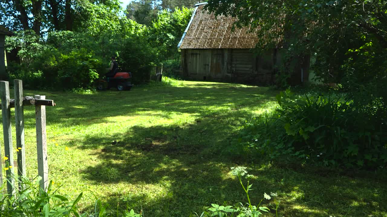 Gorgeous and colorful timelapse or time lapse footage of a man with tattoes cutting grass with a gasoline lawn mower while driving on it in front of an old vintage wooden house in the countryside.