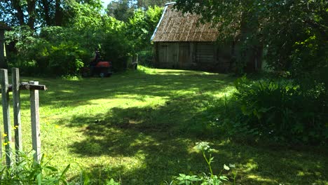 gorgeous and colorful timelapse or time lapse footage of a man with tattoes cutting grass with a gasoline lawn mower while driving on it in front of an old vintage wooden house in the countryside.