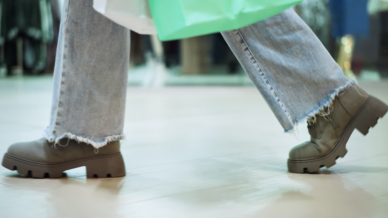 청바지 바지와 부츠를 입고 왼손으로 쇼핑 가방을 들고 쇼핑몰에서 는 개인의 다리 쪽 시각, 타일 바닥의 그림자, 배경에 흐릿한 옷