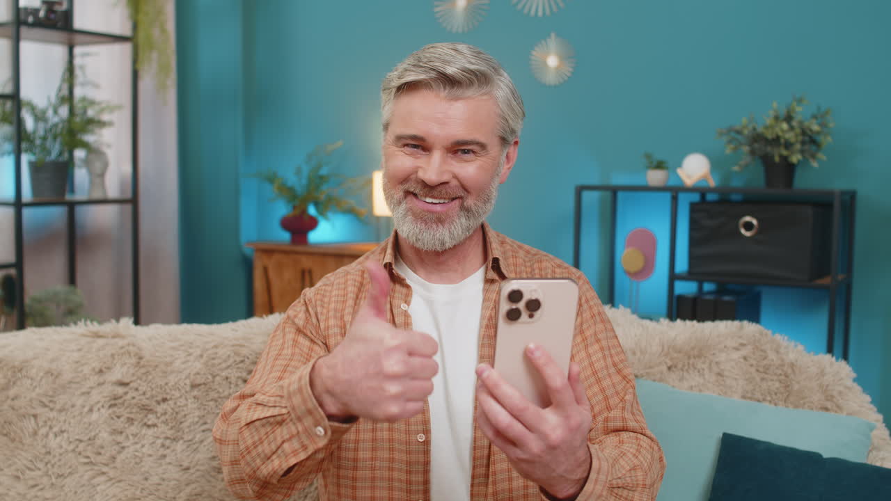 Mature man sitting on sofa with smartphone showing thumbs up like gesture on social media at home