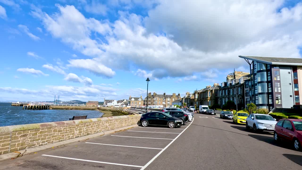 A moving camera captures parked cars, waterfront views, and modern buildings along Broughty Ferry harbour under bright daylight with scattered clouds