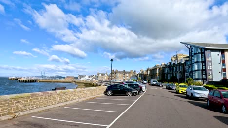 a moving camera captures parked cars, waterfront views, and modern buildings along broughty ferry harbour under bright daylight with scattered clouds