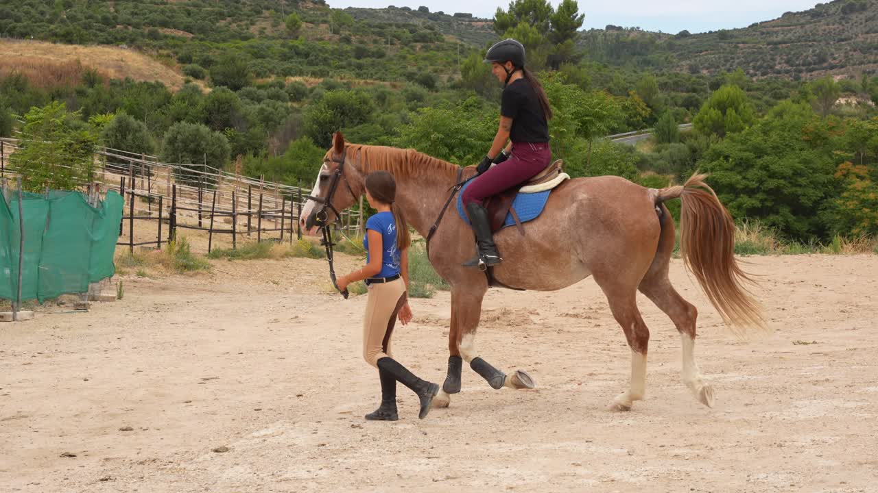 Woman instructor leads a beautiful horse with a young female rider on its back during a riding lesson in a sandy arena at an equestrian center in rural Spain