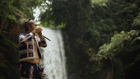 indigenous person playing kubing tribal instrument, behind is a waterfall