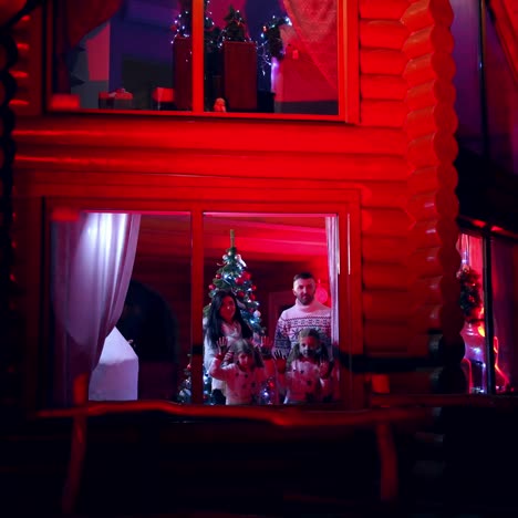 Happy young couple and kids waiting for Santa at home. Family celebrating Christmas in cosy house. Family members standing near window and looking for Christmas miracle. View from outdoors