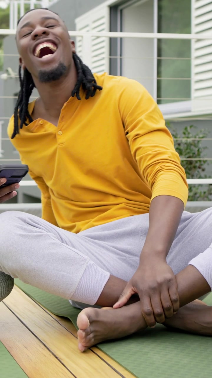 Vertical video: Adjusting foot, man in mustard top reaching phone and laughing on green mat on deck