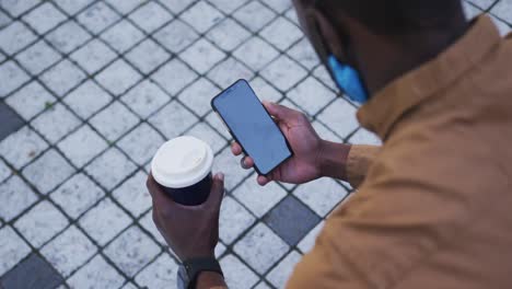 african american businessman wearing face mask using smartphone holding coffee