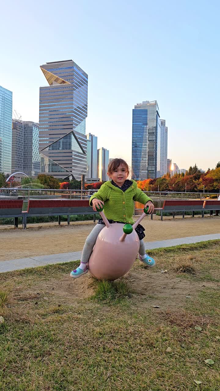 Gadis muda ceria berjaket hijau bermain di wahana patung merah muda di taman bermain Songdo Central Park dengan gedung pencakar langit modern Incheon dan dedaunan musim gugur di latar belakang