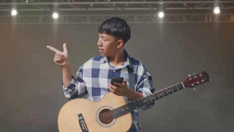 adolescente asiático con guitarra usando un teléfono inteligente y señalando al lado mientras está de pie en el escenario