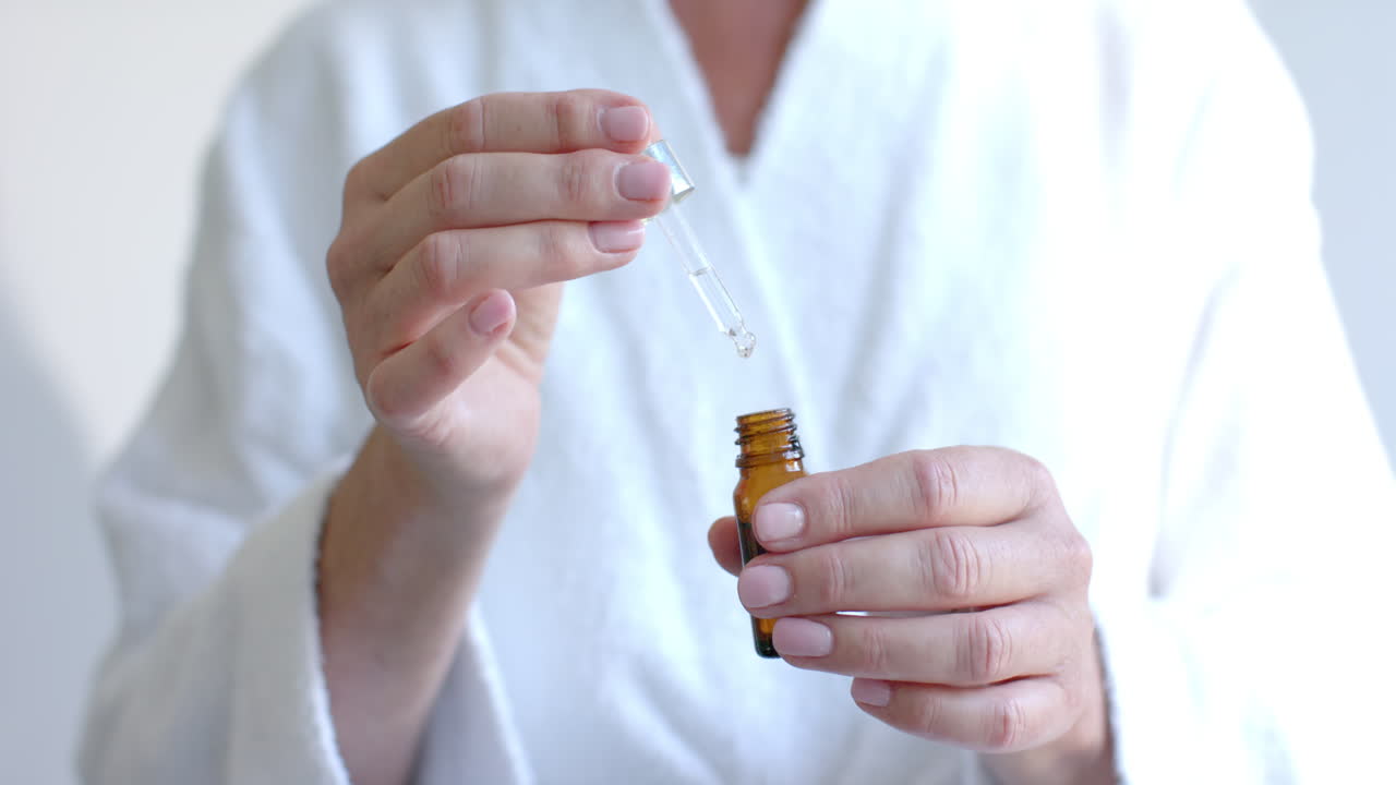 Using dropper bottle, mature woman applying skincare serum at home in bathrobe
