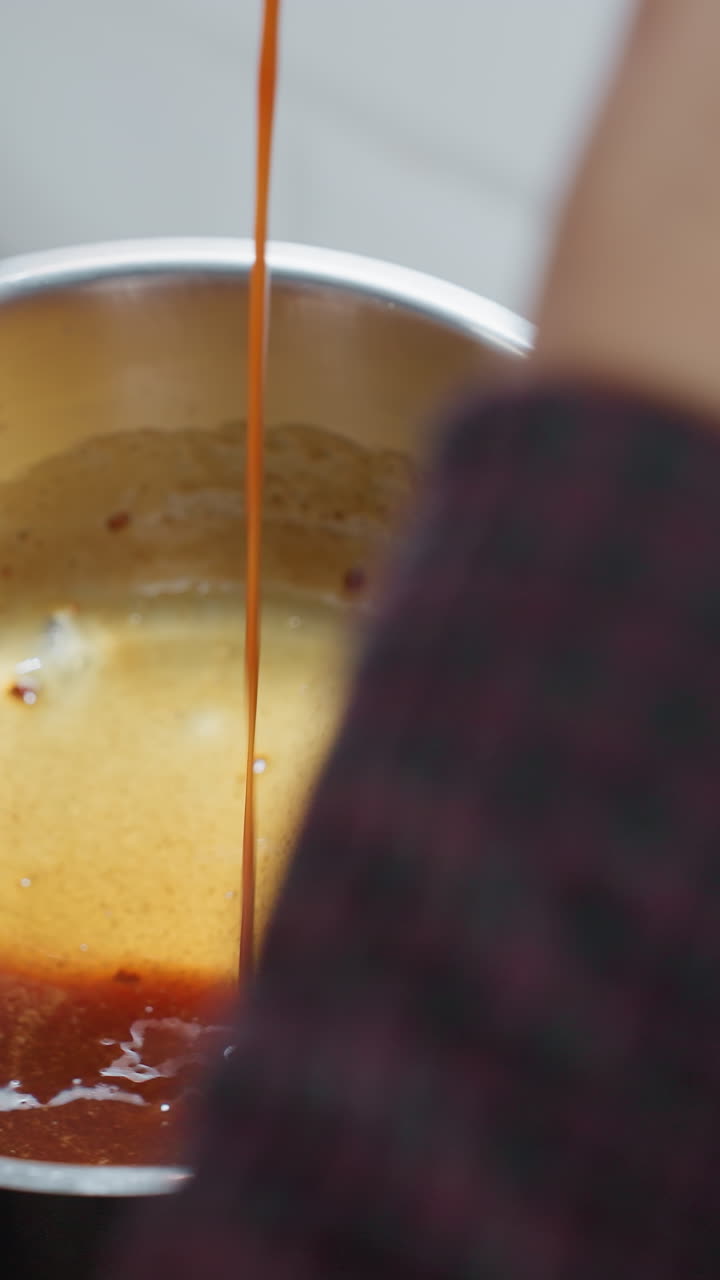 Close-up rear view of person pouring rich, simmering soup from spoon into pot in kitchen, hands with polished nails carefully handle cooking process