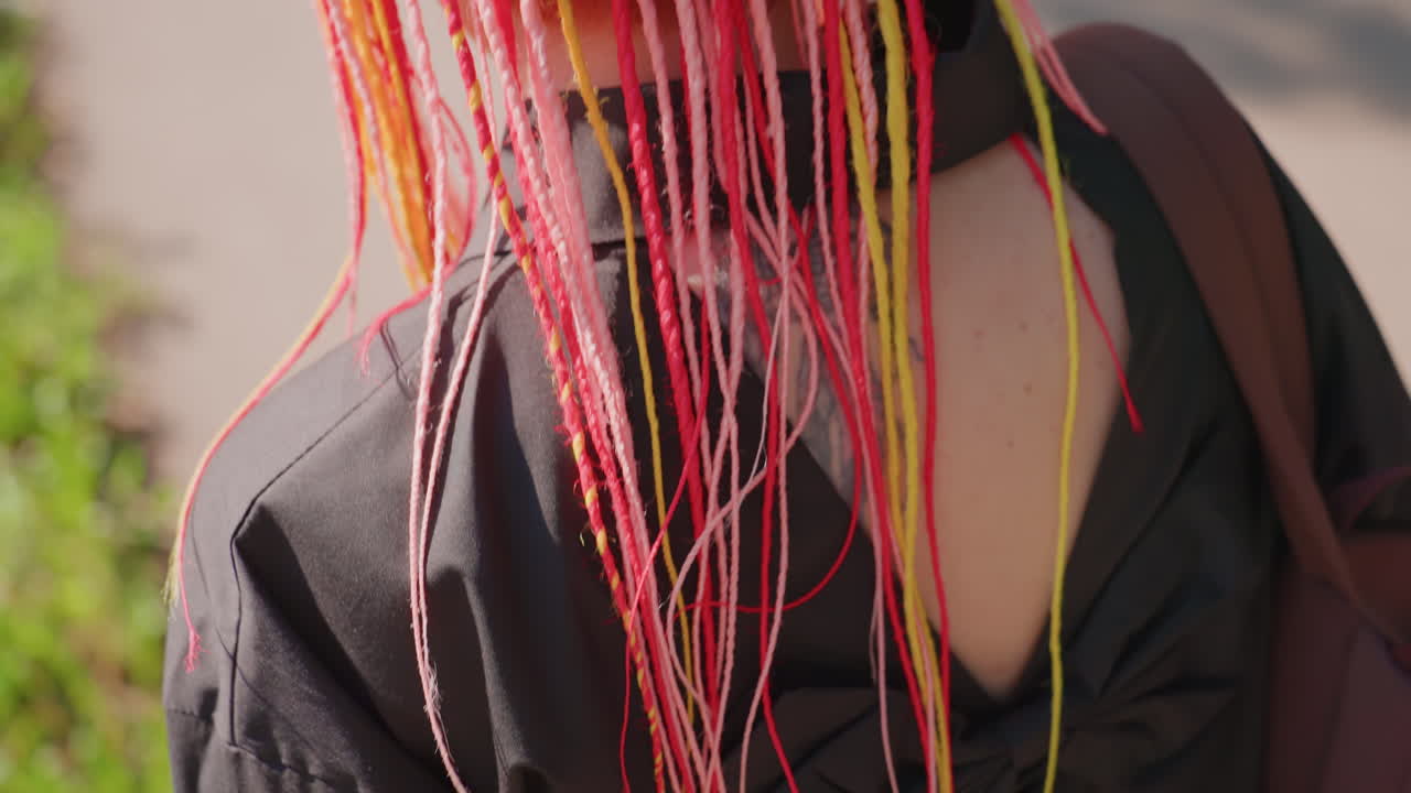 햇빛이 비치는 그림자 진 재킷 위에 다채로운 네온 색상의 머리카락이 쏟아지는 모습의 클로즈업, 햇빛이 개별 가닥을 비추며 어두운 재킷을 배경으로 흘러내리는 빛나는 네온 브레이드의 디테일 샷