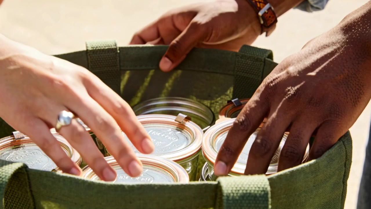 Una mano sinistra si allunga, mani con maniche arrotolate posizionano barattoli sigillati in una borsa di tela verde a terra, mettendoli al sicuro.
