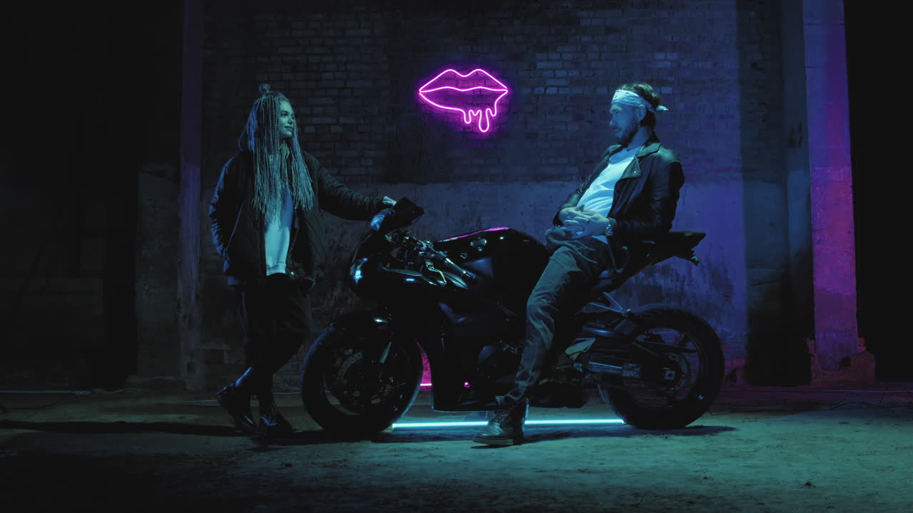 슈퍼 스포츠 오토바이를 타고 있는 한 남자가 분홍색 네온 표지판을 향해 소녀와 얘기하고 있다.