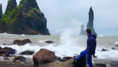 Männlicher-Tourist-Am-Schwarzen-Sandstrand-Von-Reynisfjara-In-Island,-Der-Die-Hände-Hebt,-Während-Die-Wellen-Auf-Die-Felsen-Krachen