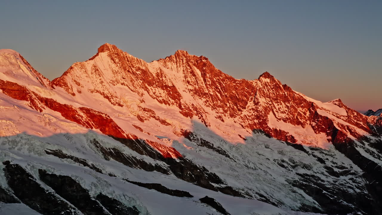 A stunning drone shot capturing the very first light of sunrise touching the Täschhorn, Dom, and Lenzspitze peaks. Golden dawn hues illuminate the towering summits of the Swiss Alps