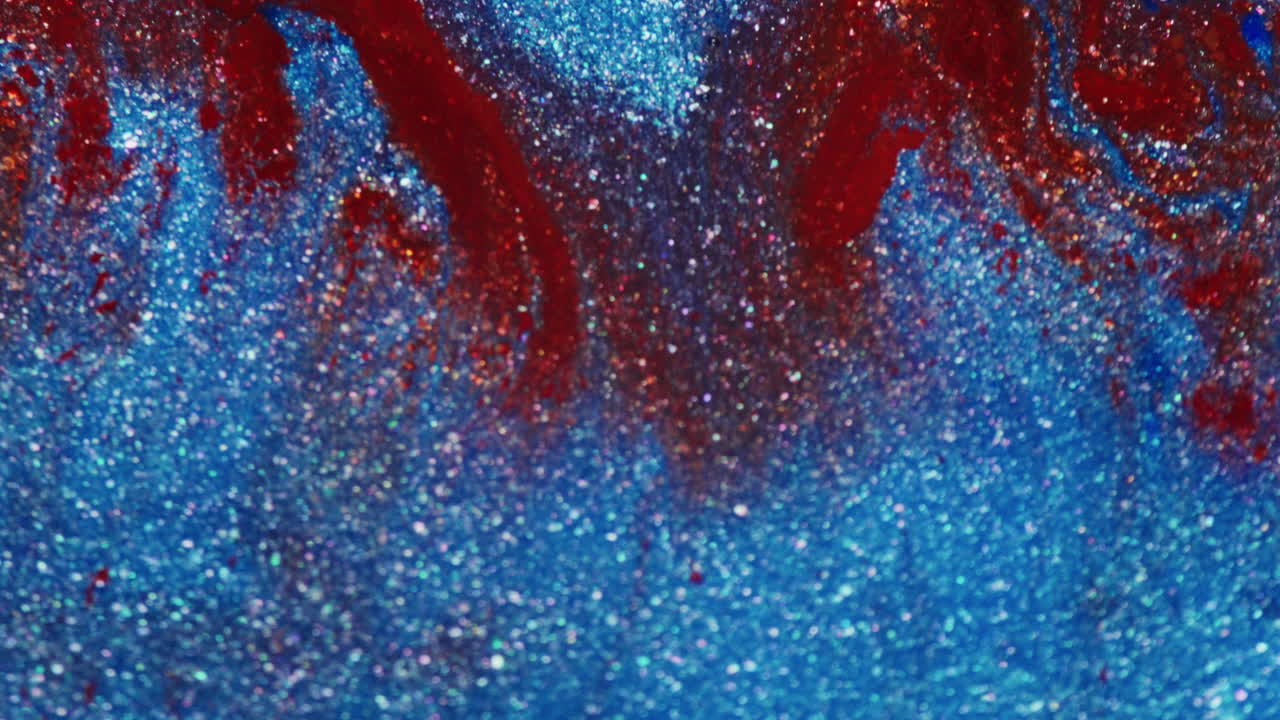 Macro shot showing red and glittery red ink being pushed into a shimmering blue glitter ink surface with visible liquid motion