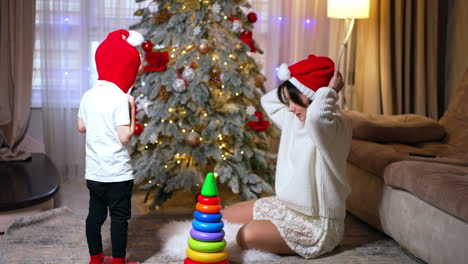 Happy young mother and little son are at home near Christmas tree. Woman and baby boy put on red Santa caps. Fun for holidays.