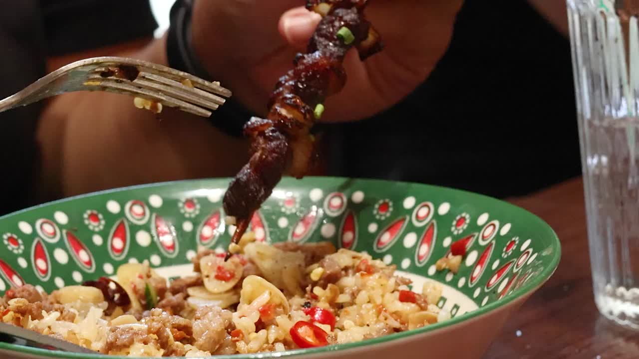 Una persona tiene uno spiedino di carne alla griglia sopra una ciotola di riso e verdure con motivi decorativi