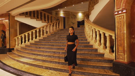 joven encantadora hermosa y elegante modelo morena caminando por lujosas escaleras interiores y dando la vuelta, vestida de negro