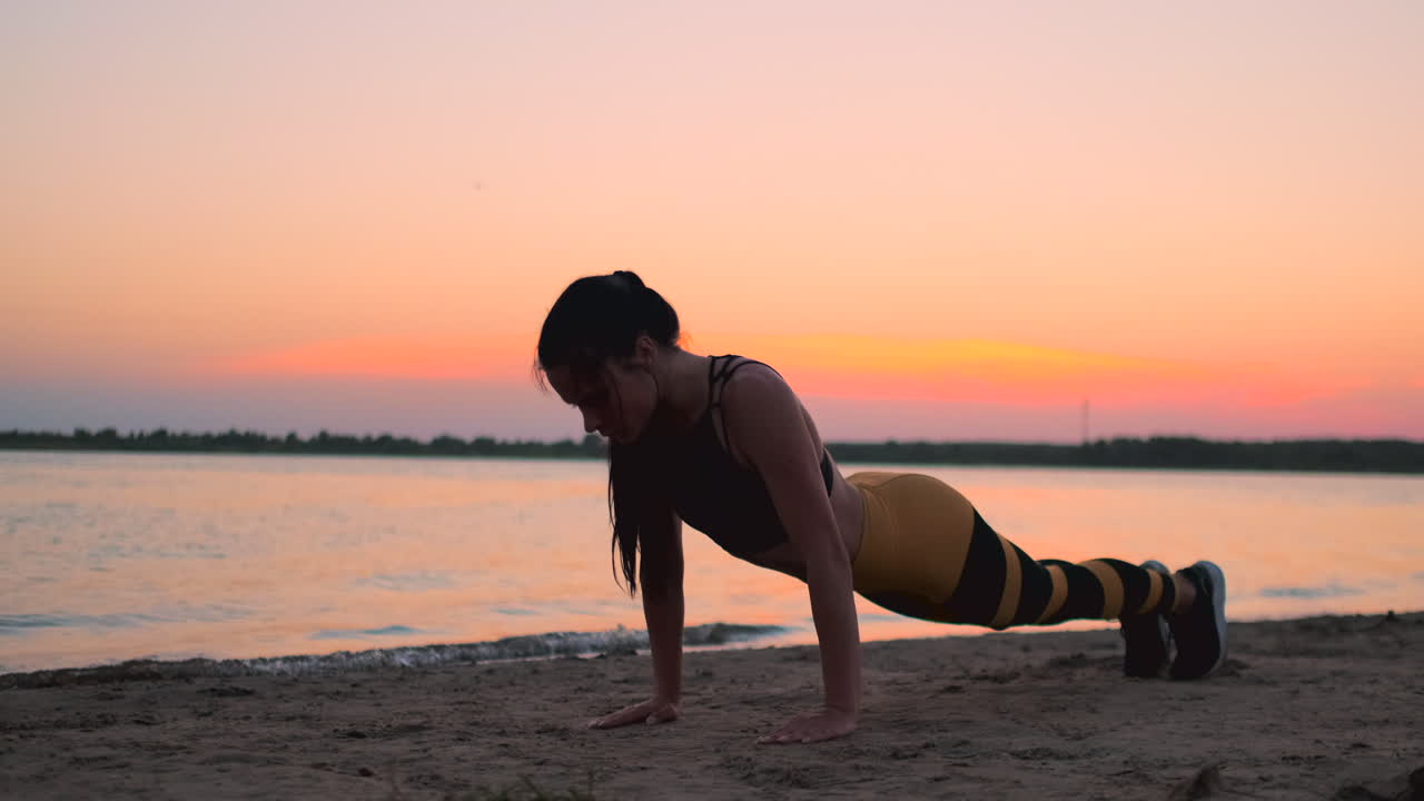 Sporty girl doing push ups on beach at sunrise. Young fit tanned smiling girl in sport top and leggings doing push ups on beach at sunset