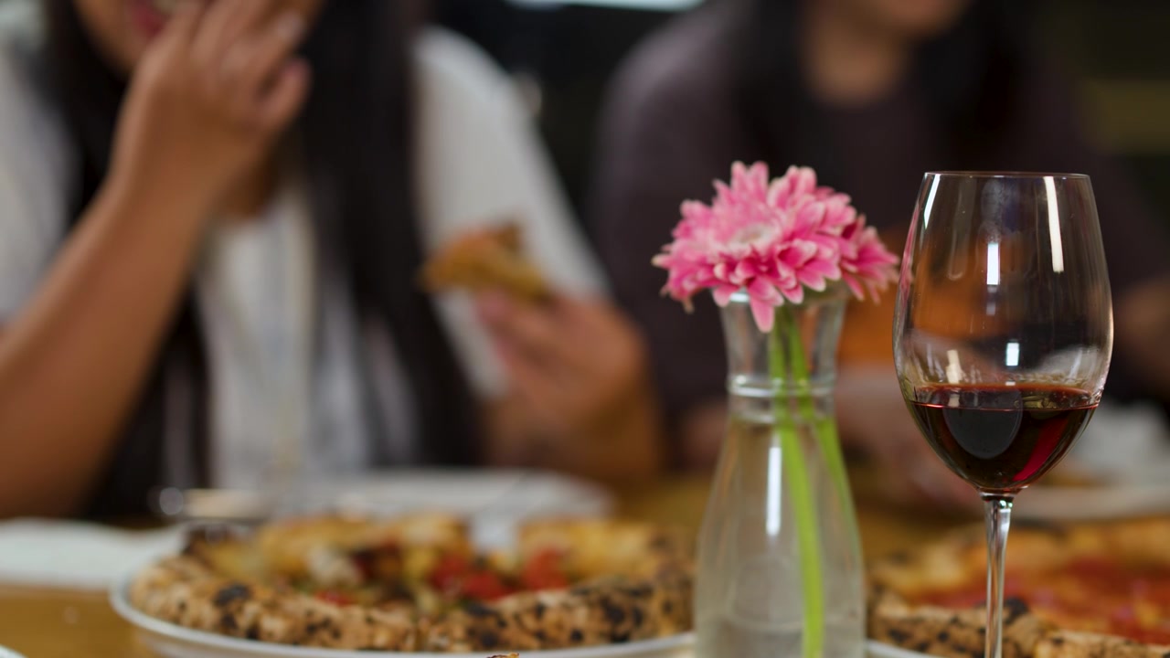 Primo piano di pizza e vino su un tavolo, mentre gli amici mangiano sullo sfondo sfocato.