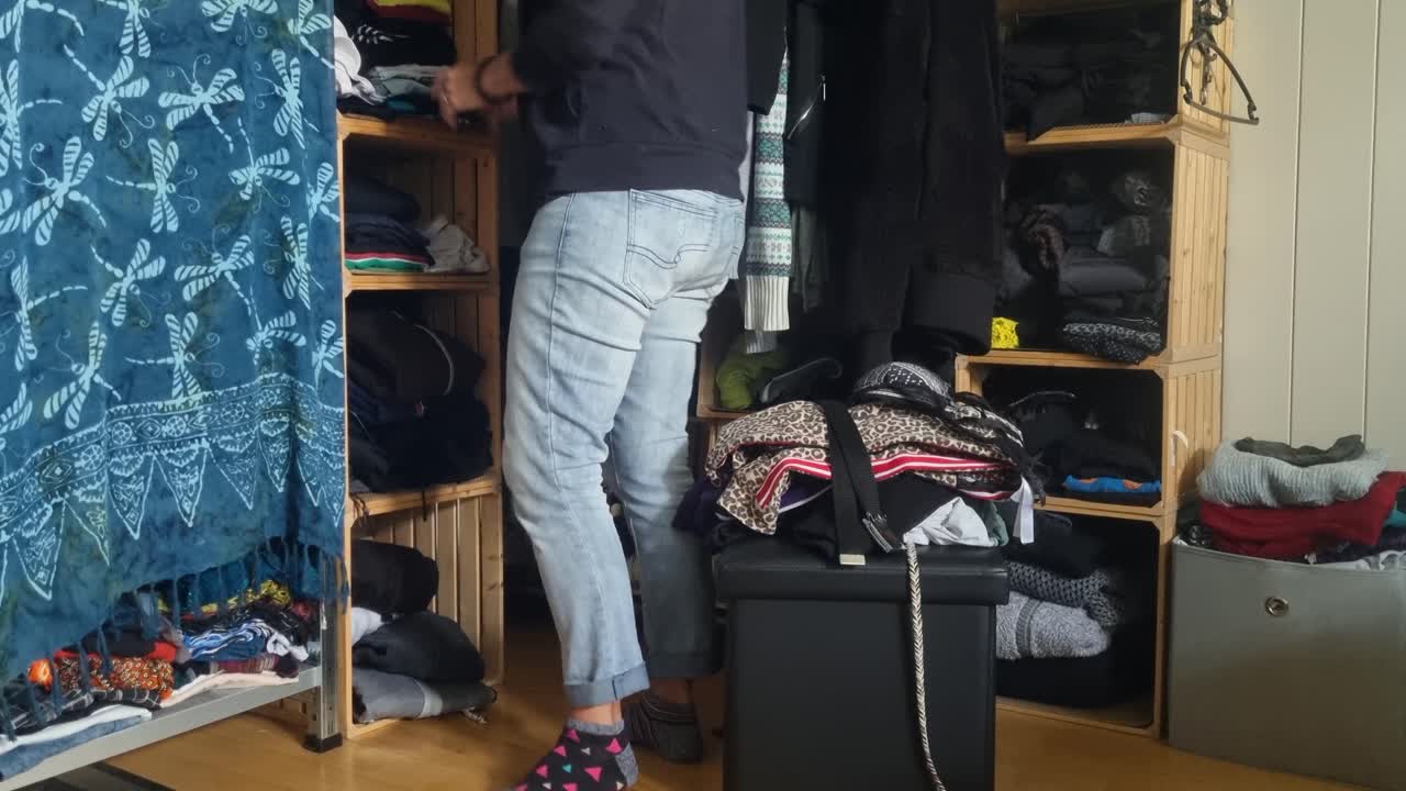 A woman in jeans sorts through clothes in her messy open closet, walking around her home in Switzerland and trying to organize her wardrobe