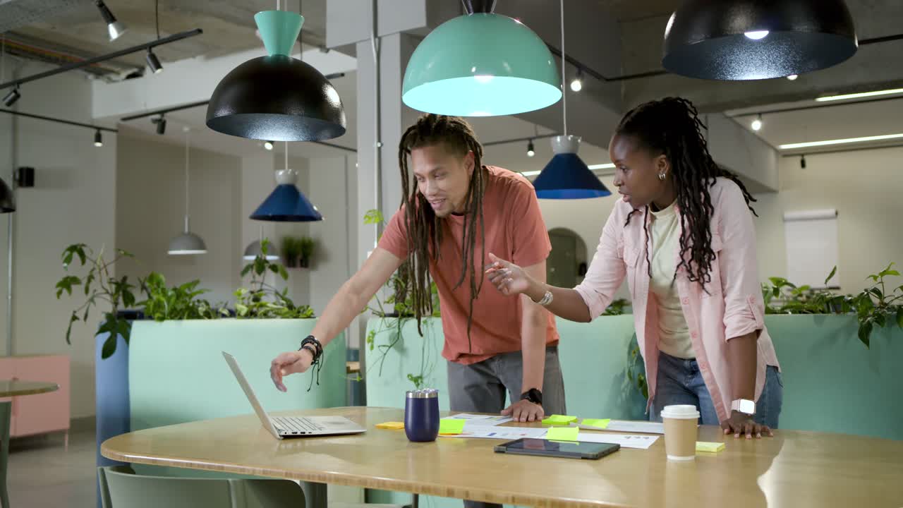 African American team leaning at table, man pointing pen at laptop urging woman sorting neon notes