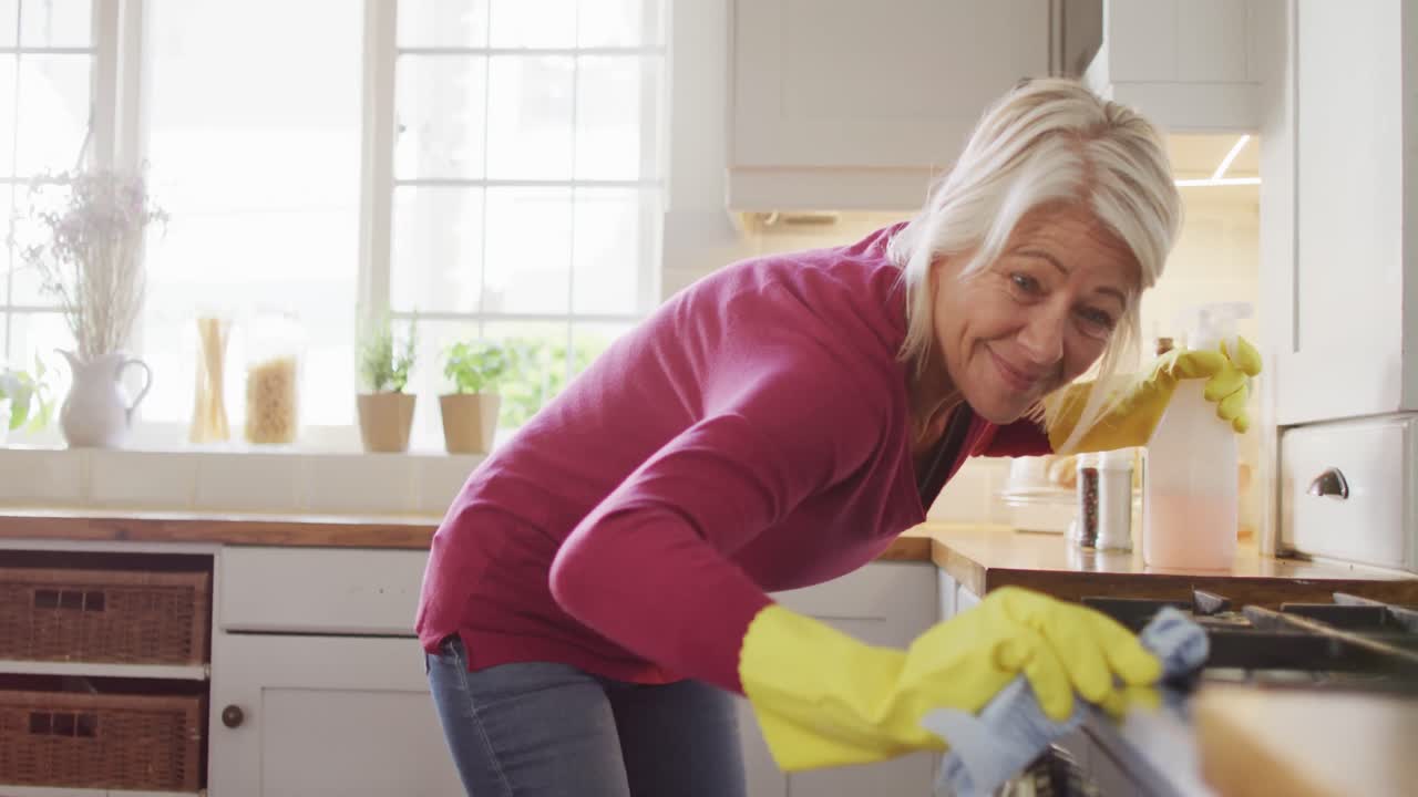 mujer caucásica mayor feliz usando guantes de goma, limpiando la encimera en la cocina