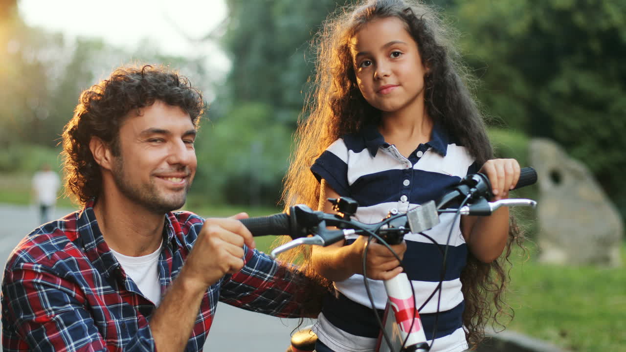 Closeup. Portrait of a little girl and her dad next to the bike. Dad hugs her. Looking at each other, then - into the camera. Smiling. Blurred background