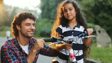 Closeup.-Portrait-of-a-little-girl-and-her-dad-next-to-the-bike.-Dad-hugs-her.-Looking-at-each-other,-then---into-the-camera.-Smiling.-Blurred-background