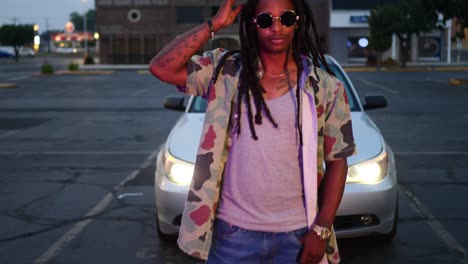 african american man with dreadlocks wearing dark circle sunglasses in front of car headlights and adjusting his hair as camera moves in towards him