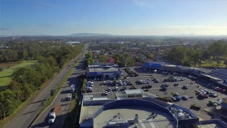 toma aérea de un dron pasando por un centro comercial local hacia una concurrida vía arterial, con el monte coot-tha y el centro de brisbane en la distancia