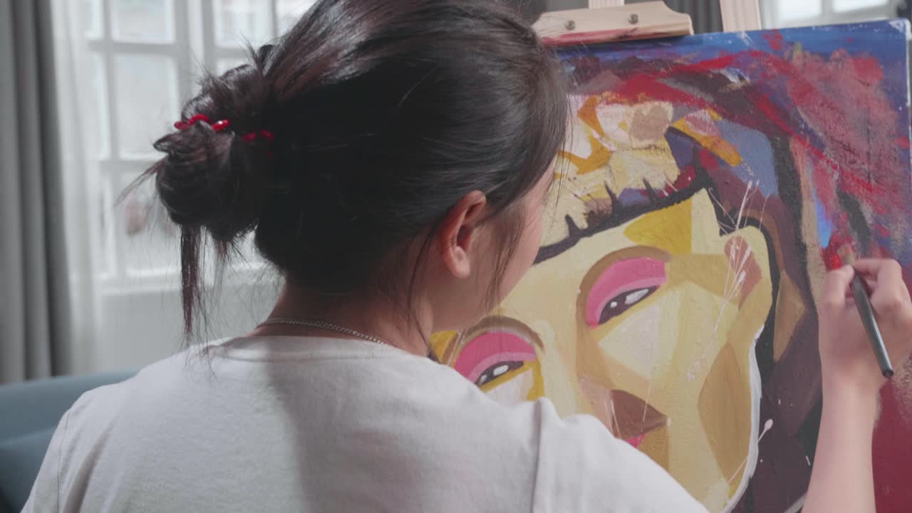 Back View Of An Asian Artist Girl In Wheelchair Holding Paintbrush Mixed Colour And Painting A Girl On The Canvas
