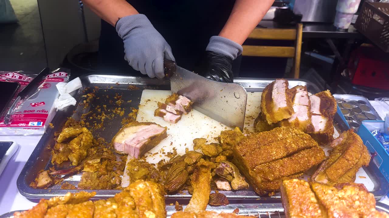A street vendor expertly slices crispy pork under bright lighting in a bustling Bangkok market, showcasing culinary skill and vibrant street food culture