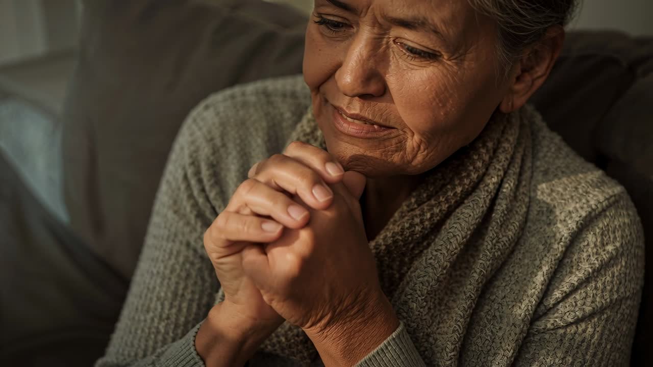 Sintiendo rigidez, una mujer mayor se frota y masajea las manos en el sofá de casa, vistiendo un suéter gris.