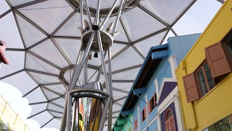vibrant, multi-colored shophouses line a pedestrian street beneath a geometric canopy in singapore. daylight illuminates the urban scene as the camera tilts upward