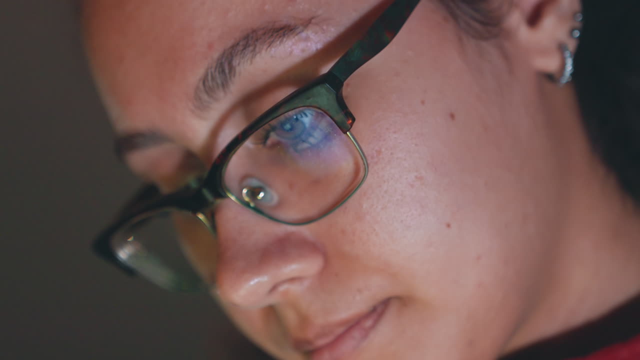 Primo piano di una donna con occhiali che studia appunti di ricerca sullo schermo, con un sottile bagliore blu riflesso nelle lenti. Ha un'espressione quieta e concentrata, con piercing all'orecchio visibili, illuminata da una morbida luce serale, che trasmette una profonda concentrazione accademica.