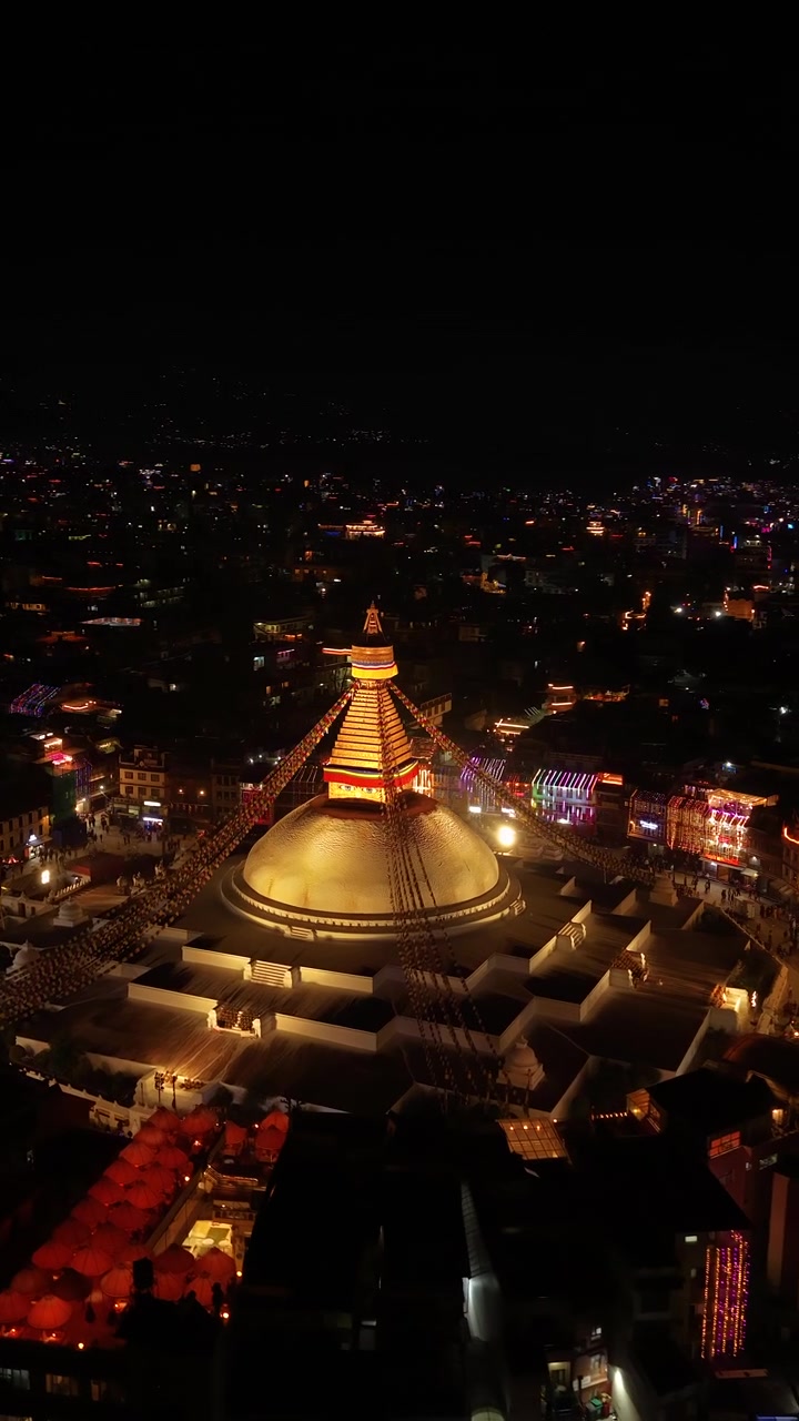 Kathmandu, Nepal, inquadratura aerea verticale 9:16 che si avvicina alla cupola dorata splendidamente illuminata della Stupa di Boudhanath, circondata da luci colorate della città durante la notte del festival Tihar