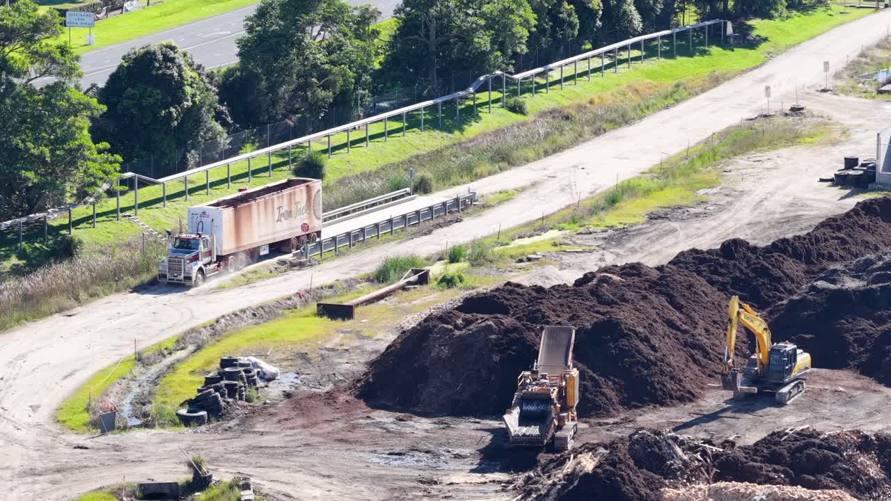 Drone footage captures an excavator loading soil onto a truck in a lush, industrial setting under bright daylight