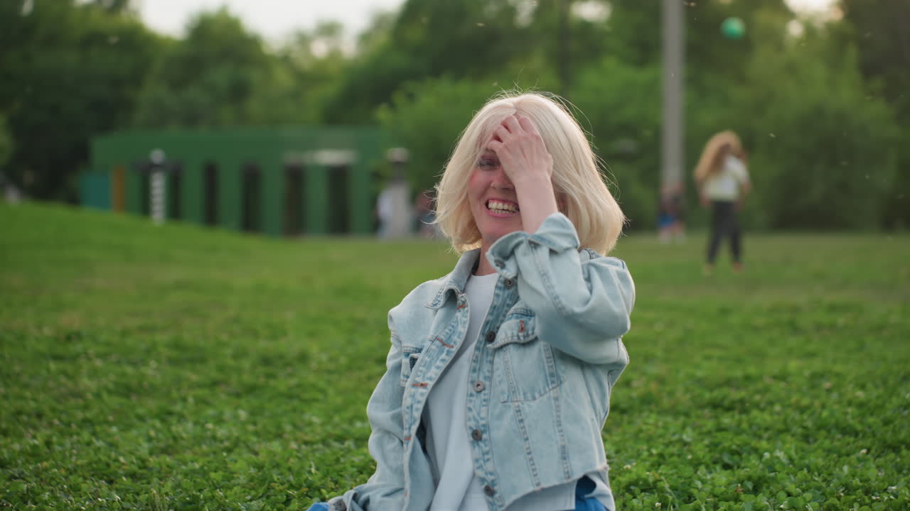 young woman sitting on grass laughing loudly while watching people in park, casual denim jacket and blue pants, blurred figures playing nearby, summer evening light creating carefree joyful mood