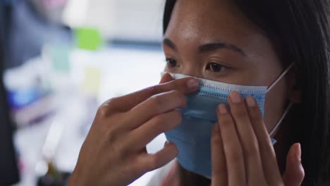 Mujer-Asiática-Ajustando-Su-Mascarilla-Mientras-Está-Sentada-En-Su-Escritorio-En-La-Oficina-Moderna