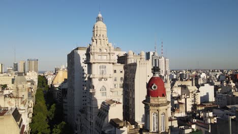 Aerial-rising-over-Barolo-palace-tower-in-tree-lined-May-Avenue-surrounded-by-Monserrat-buildings-at-sunset,-Buenos-Aires