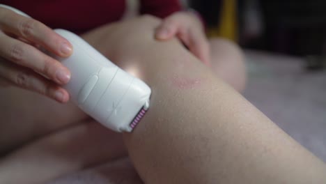 mujer que usa una herramienta depilatoria de maquinilla de afeitar eléctrica con luz en la pierna para eliminar el vello mientras se sienta en la cama