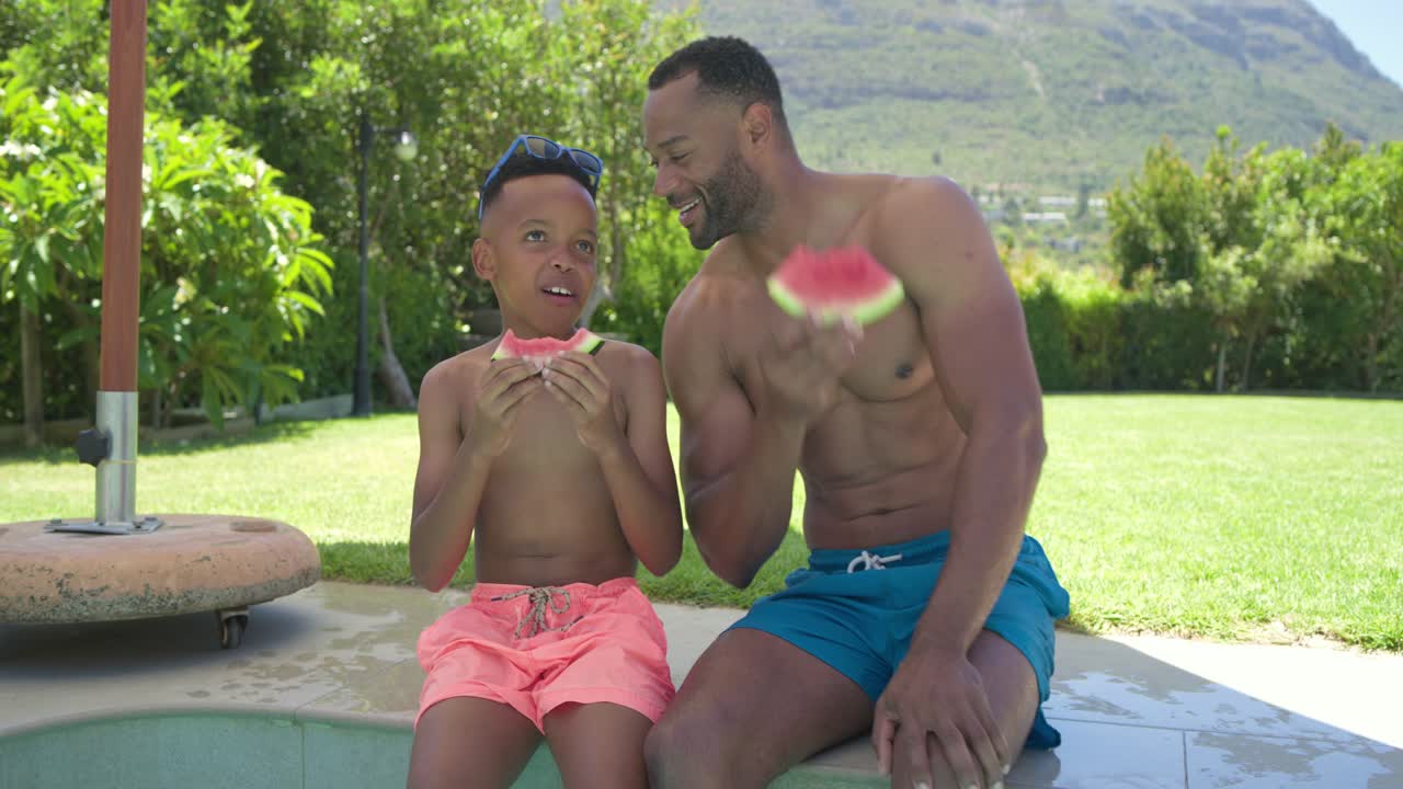 アフリカ系アメリカ人の父親と若い息子がプールサイドに座り、父親がスイカを掲げ、息子は笑いながら絆を深めている様子。