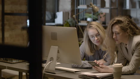 two female employees looking something at mobile phone and talking together while working in the office