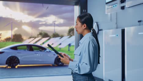 side view of asian female professional worker with home energy storage system in a modern garage, observes by looking up before she come to concentrating on the tablet and keep on checking